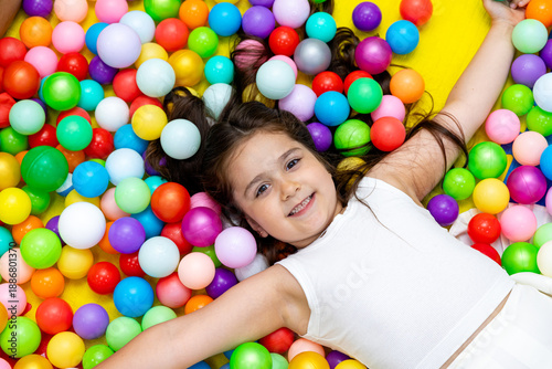 Cute little girl lying on multi coloured plastic balls in big dry paddling pool in playing centre. Smiling at camera. Portrait close up. Having fun in playroom. Leisure Activity.