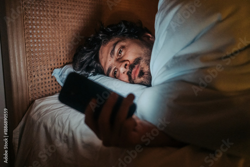Bild auf Leinwand Man lying in bed at night, looking at his smartphone, suggesting themes of sleeplessness, technology use in the bedroom, or potential negative impact
