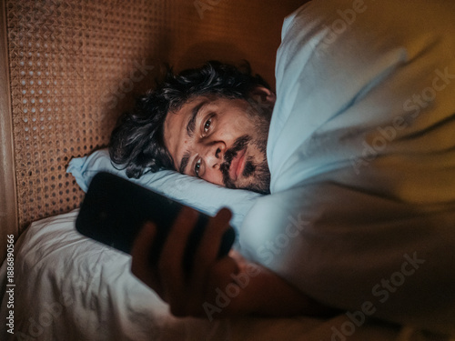 Man lying in bed at night, looking at his smartphone, suggesting themes of sleeplessness, technology use in the bedroom, or potential negative impact. 