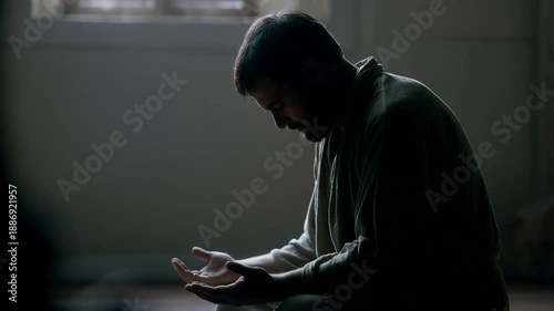 A man in prayer, kneeling in a dimly lit room with his hands cupped in front of him, with a window in the background