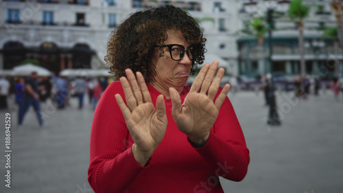Foto Woman in red sweater raising hands and wrinkling nose in crowded street plaza with blurred pedestrians; disgust