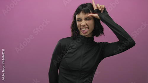 Canvas Print Woman forms loser sign on forehead in pink studio setting with scrunched face and tongue out; playful defiance