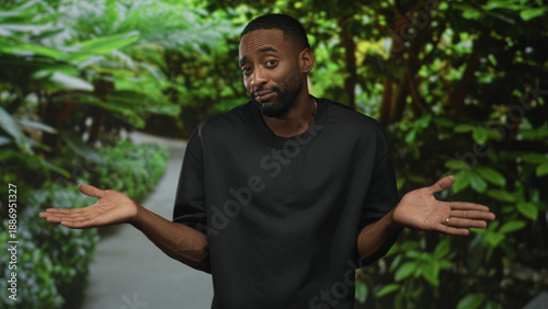 Man in black t shirt shrugs with palms up on a leafy green park path, facing camera with a slight smile; uncertainty casual relaxed.