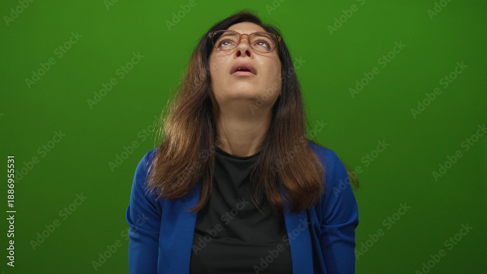 Fototapeta premium Hispanic woman raising hands toward ceiling and tilting head back in wide green studio set; frustration.