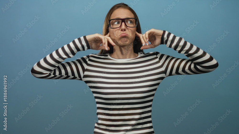 Fototapeta premium Woman in striped shirt and glasses plugs ears with fingers in blue studio setting while squinting and pursing lips; frustration defiance.
