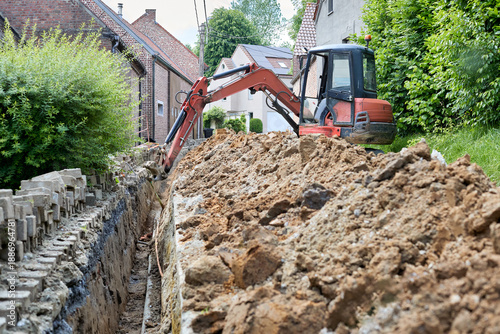 Canvastavla A small orange excavator digs a deep trench through an urban area for utility installation