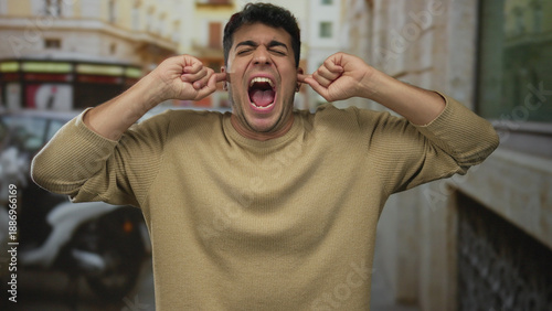Man presses ears to cover blaring street horns as hispanic male stands outdoor amid noise with rising annoyance and discomfort.