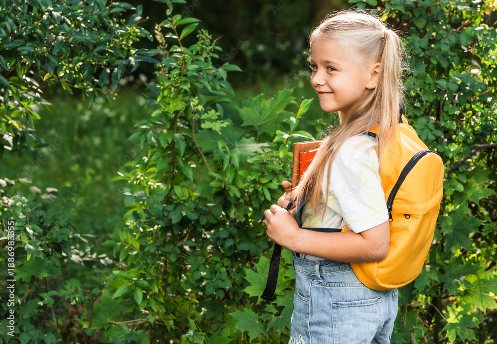 Fototapeta premium Child girl with yellow backpack in the green school yard.