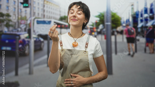 Valokuva Woman jeweler holding a small ring between fingers on a city street, wearing apron and orange pendant while smiling; craft pride
