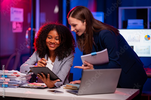 Obraz na plátně Two cheerful businesswomen are working intensely together, analyzing data and exchanging ideas to explain business reports to colleagues while working overtime late into the night
