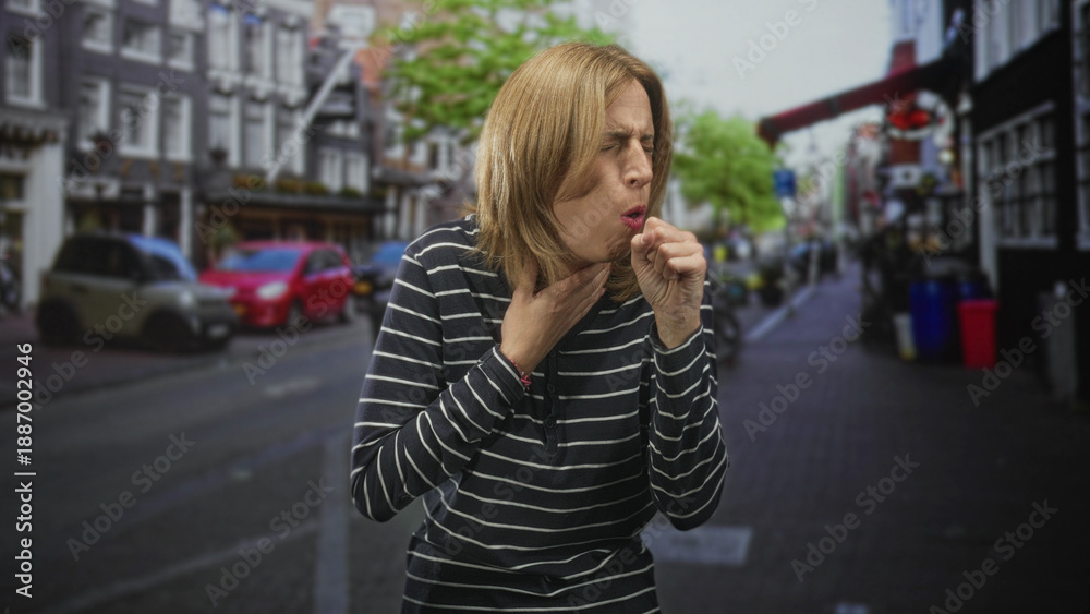 Fototapeta premium Woman coughing into fist with hand on throat on a city street, striped top visible; illness discomfort.