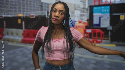 Fotografija Woman with crossed arms showing exposed arms and stern face in a pink crop top on street with construction barriers and worker visible; defiance resilience