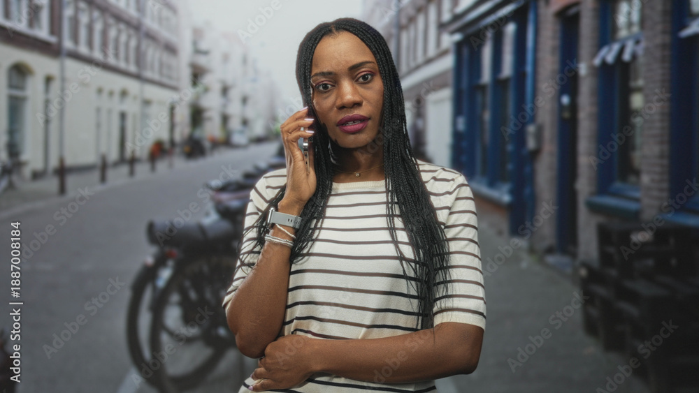 Fototapeta premium Woman holding smartphone, looking at screen with hand visible, wearing striped top and bracelets on a narrow city street near bicycles and storefronts; quiet concern.