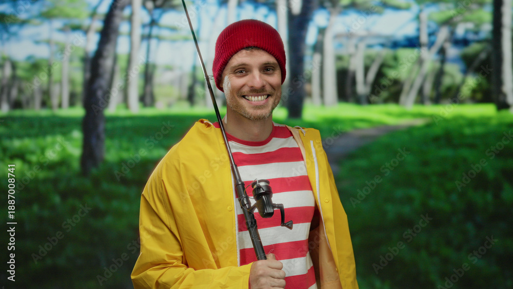 Naklejka premium Young man smiling in a vibrant park setting, wearing a red beanie and yellow jacket, holding a fishing rod with lush green trees in the background.