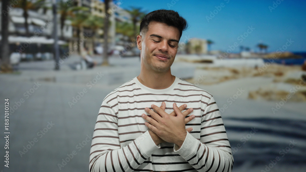 © Krakenimages.com - Young man in striped shirt at beach expressing contentment with eyes closed, hands on chest, outdoors beside the ocean, amid coastal landscape and palm trees