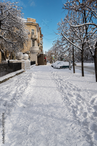 Winter city street. Roads, sidewalks, and parked cars are covered with snow. A sunny January day. After a heavy overnight snowfall. New Year's Morning. Old houses. The road is almost empty.