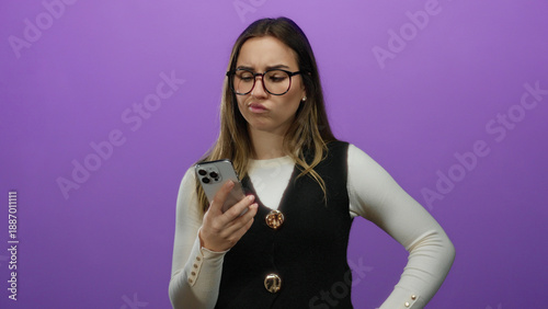 Принти на полотні Young woman wearing glasses looks puzzled while holding smartphone against isola