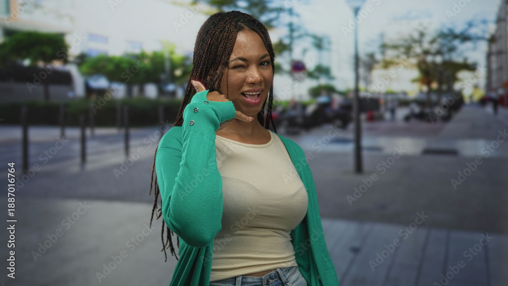 Fototapeta premium Young african american woman smiling and showing call gesture with hand and wink on a busy urban street; playful confidence.