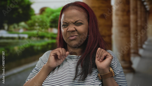 Obraz na plátně Woman with red hair recoils with clenched fists on sunny street lined with stone columns wearing striped shirt; disgust
