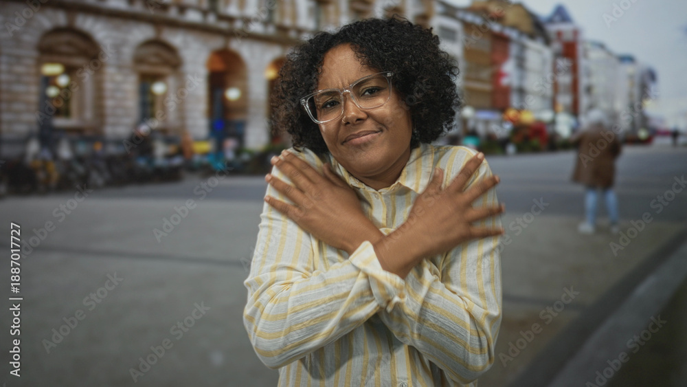 Fototapeta premium Young african american woman wearing glasses crosses arms on street lined with stone buildings; anxiety.