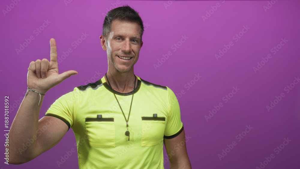 Fototapeta premium Man referee pointing index finger upward while smiling, wearing yellow referee shirt and whistle in purple studio; confidence authority.
