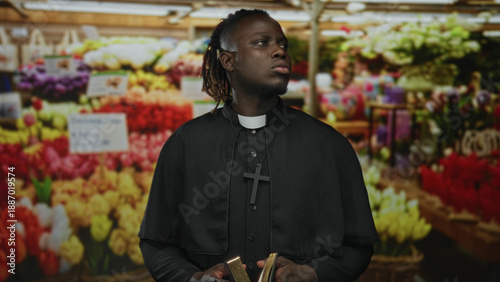 Картината върху платно Young priest man holding book with visible hands and clerical collar in flower market stalls; faith serenity