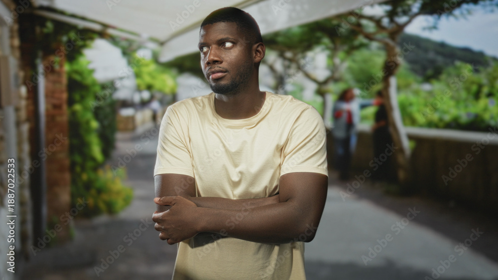Fototapeta premium Man with folded arms holding his elbow, standing on a street under a canopy by trees and a stone railing; pensive solitude.