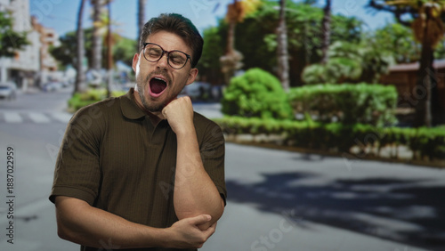 Man yawning with hand to mouth on city street near palm trees and crosswalk, wearing glasses and polo shirt; sleepiness commute.