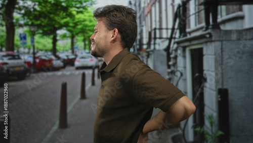 Wallpaper Mural Man holding lower back and leaning forward slightly, hand at waist showing backache on street with parked cars and bollards; frustration. Torontodigital.ca