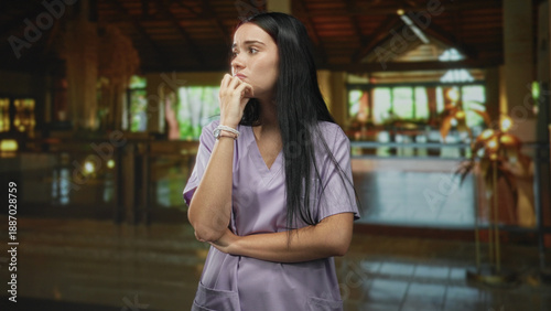 Bilde på lerret Woman nurse in lavender scrubs with hand to mouth and crossed arm, standing in a building lobby; quiet concern