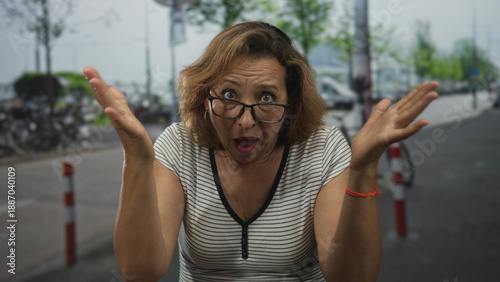 Foto Middle age hispanic woman in striped shirt adjusts glasses on city street lined with parked bicycles and bollards; surprise curiosity
