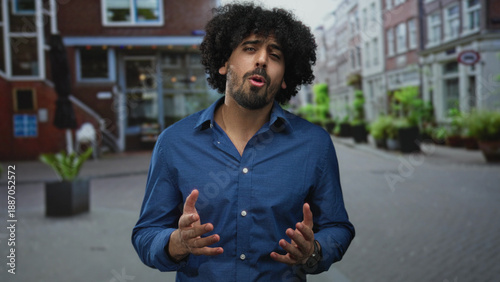 Photos Man with curly hair and visible hands gesturing while speaking on a city street in a blue shirt; confidence persuasive conversation