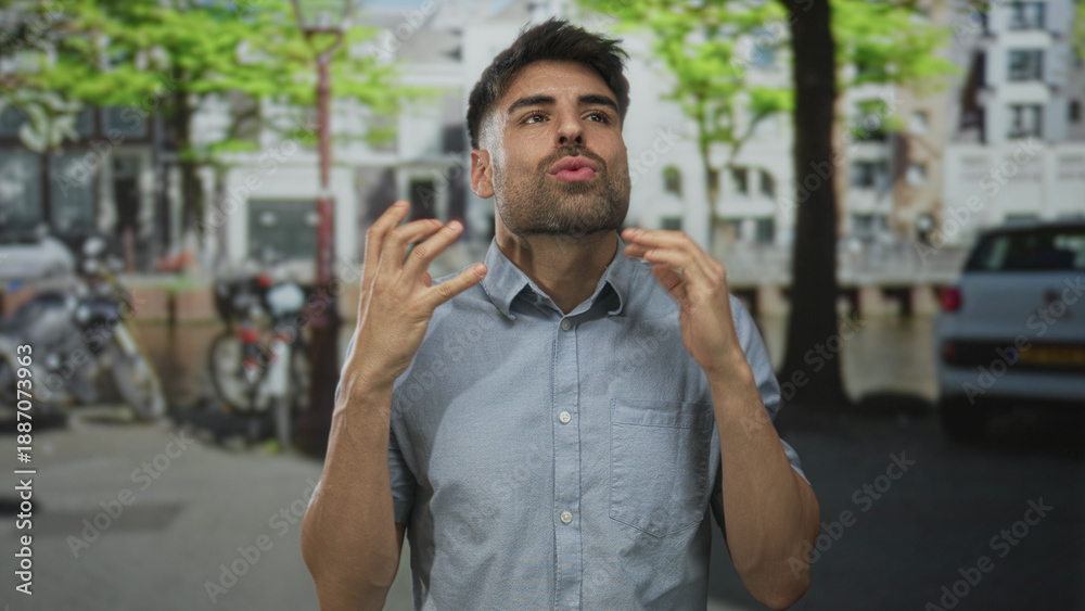 Obraz premium Man raising hands by canal on bustling street lined with parked bikes and cars under green trees; frustration.