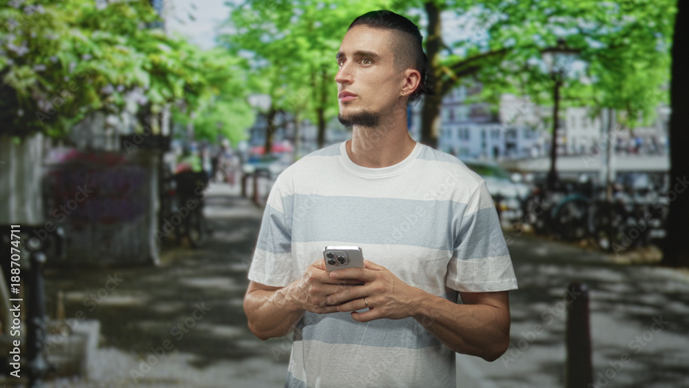 Fototapeta premium Man holding smartphone with both hands and looking aside on a tree lined city street with bicycles and parked cars; contemplation urban life.