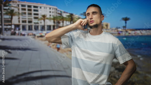 Man holding smartphone to ear, talking on street promenade by the sea with buildings and palm trees visible; quiet contemplation. Free Download Man holding smartphone to ear, talking on street promenade by the sea with buildings and palm trees visible; quiet contemplation. - GFXLoad