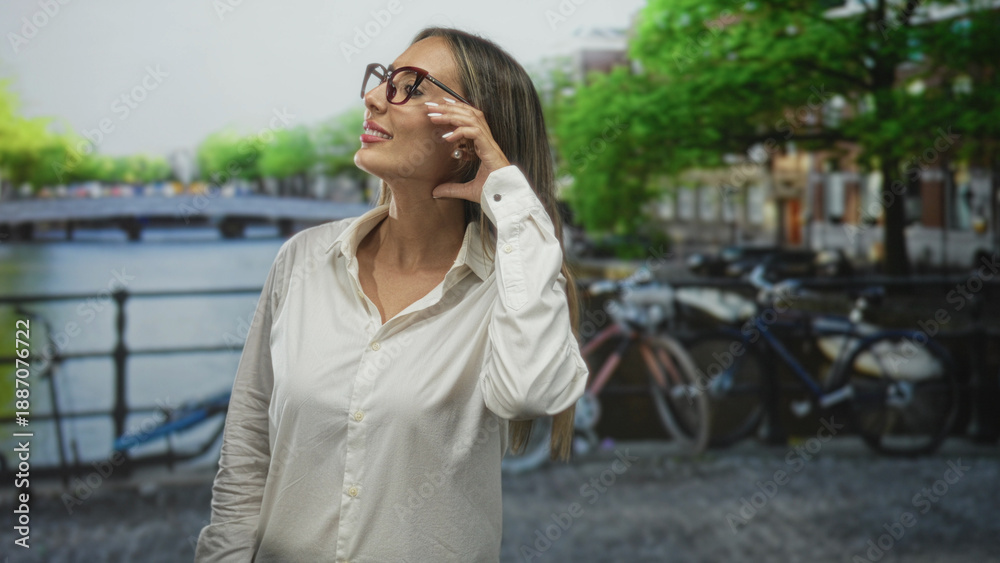 Fototapeta premium Woman tucks hair behind ear while adjusting glasses on canal street in amsterdam, near bikes and water; calm reflection.