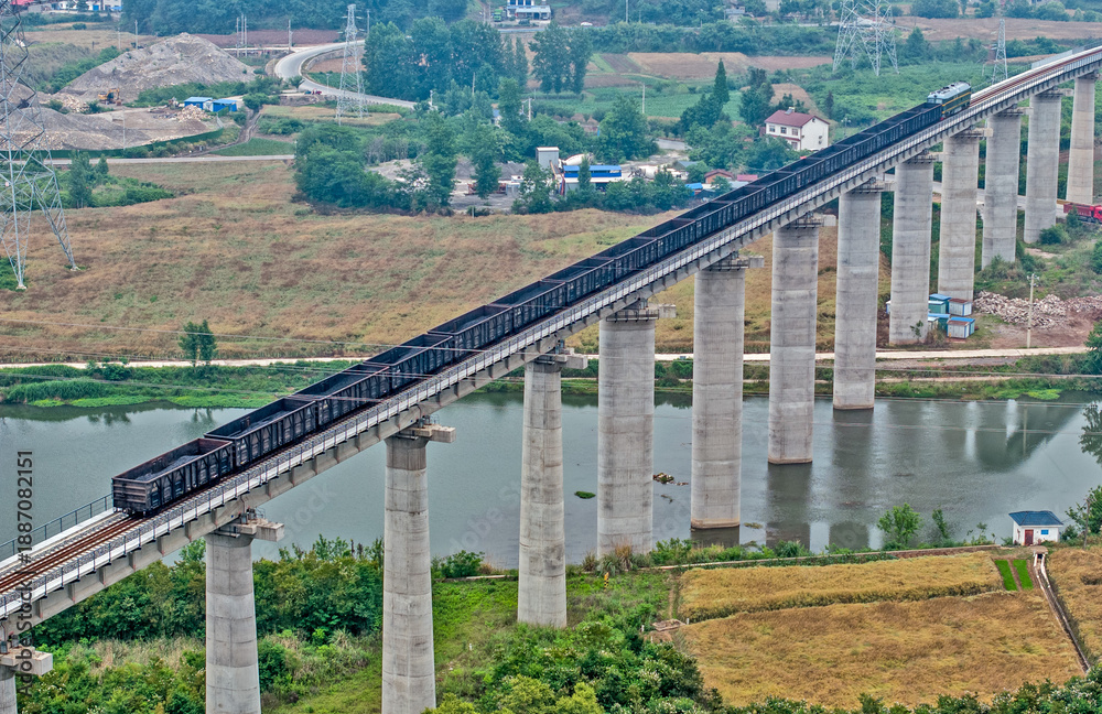 Obraz premium Freight Train on High Viaduct Crossing River Through Rural Landscape