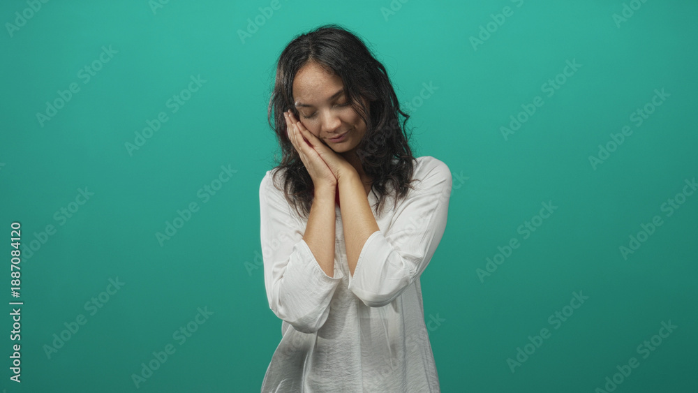 Obraz premium Young hispanic woman resting hands against cheek in a teal studio with white blouse and soft lighting, gentle pose and closed eyes; peaceful rest.
