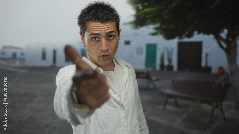 Fototapeta premium Young hispanic man wearing loose white shirt points finger to camera on street showing hoop earrings; anger frustration tension confrontation.