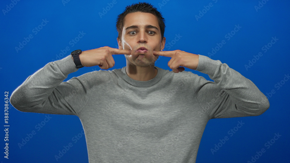 Naklejka premium Young hispanic man making funny face in grey sweater against isolated blue background wall with fingers pointing at cheeks for humorous expression
