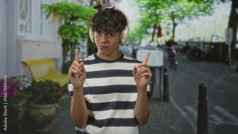 Fototapeta premium Man wearing headphones points both index fingers while standing on a street by a bench with potted flowers; playful listening.