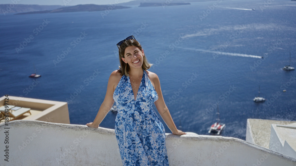 Fototapeta premium Woman smiling with arms out on building terrace overlooking sea and boats in santorini town; vacation relaxation joy.