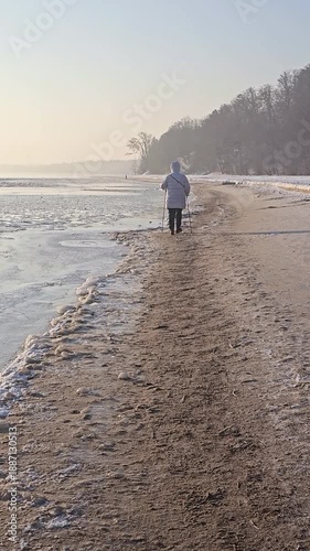 Person old woman practicing Nordic walking along the seaside in winter, combining fitness and nature in a cold coastal setting.