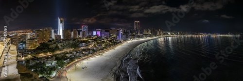 Elevated view from Mirador de l'Ermita Verge del Mar in Benidorm city by night, with city shoreline, Poniente beach and a calm sea, road and buildings enlightened under a cloudy dark sky 