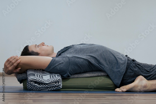 Фотография Young adult man lays on yoga bolster stretching his spine and hips, face up at r