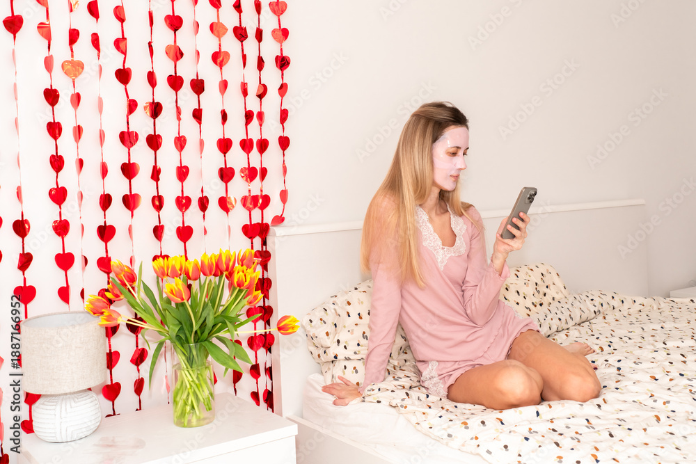 Obraz premium Woman in sheet face mask relaxing on bed while using smartphone. Home spa atmosphere, beauty care, wellness lifestyle and modern self care concept