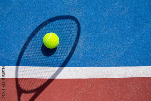 Tennis ball on a blue outdoor court with a visible racket shadow.
