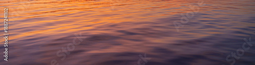 Fotografi Golden Hour Reflections on Calm Water Surface,Abstract nature background of gentle ripples on a water surface during sunset, featuring a vibrant blend of golden orange and deep purple hues