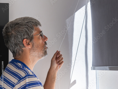 Bild auf Leinwand Man with gray hair and glasses opening a window roller blind, starting the morni