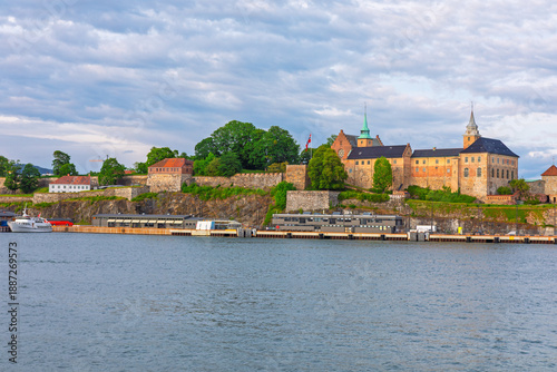 Akershus Fortress situated on headland overlooking the calm waters of Oslofjord in Oslo, Norway. Medieval fortress complex is surrounded by lush trees and fortified walls with boat dock at its base